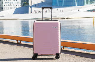 Rosa hardplastkoffert med fire hjul ved vannkanten.