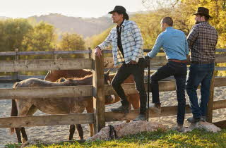 Tre menn i jeans, rutete skjorter og cowboy hatter sår og lener seg mot en innhegning med hester.