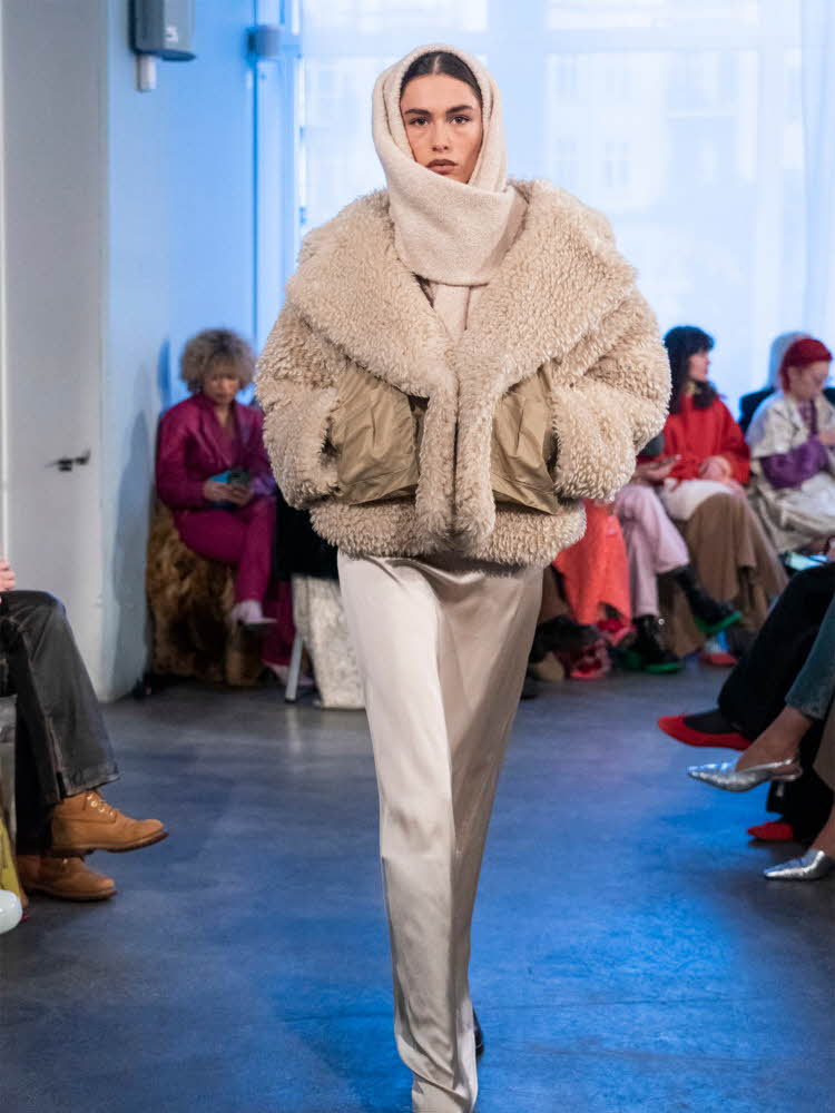 Damemodell på catwalk med skjerf rundt hodet, teddyjakke, hvitt skjørt og sorte sko.