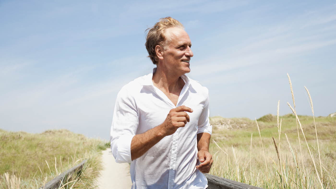 Eldre mann på strand iført hvit skjorte. Sand og gress og blå himmel.