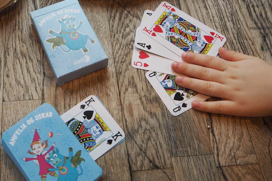 Amfelia og Oskar-kortstokk på et trebord, samt en barnehånd som holder på kortene