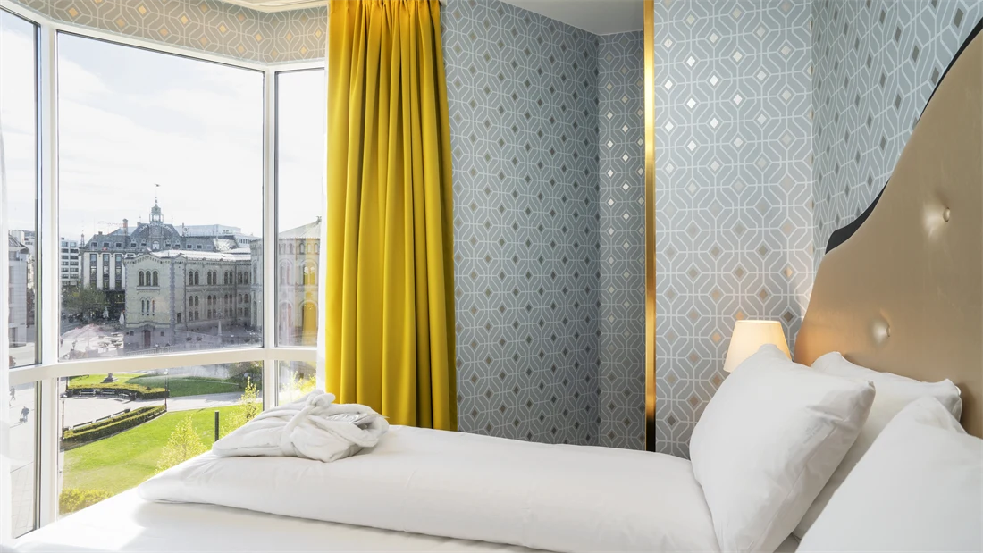 Dobbeltseng, mønsteret sølv tapet, gule gardiner og store vinduer med utsikt over Stortinget i junior suite på Thon Hotel Cecil