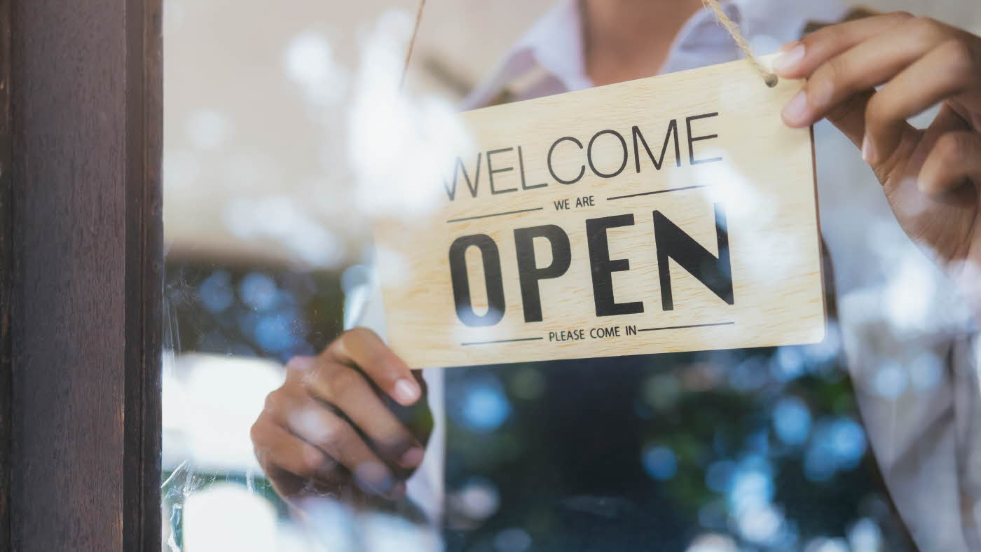 Butikkansatt på innside av glassdør som holder et skilt det står "Welcome - we are open - please come in" på.