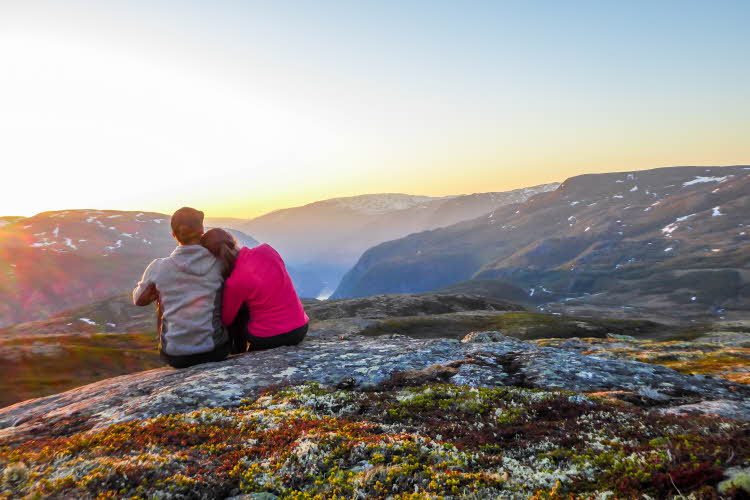 To mennesker sitter på en fjelltopp og skuer utover fjellheimen i solnedgang