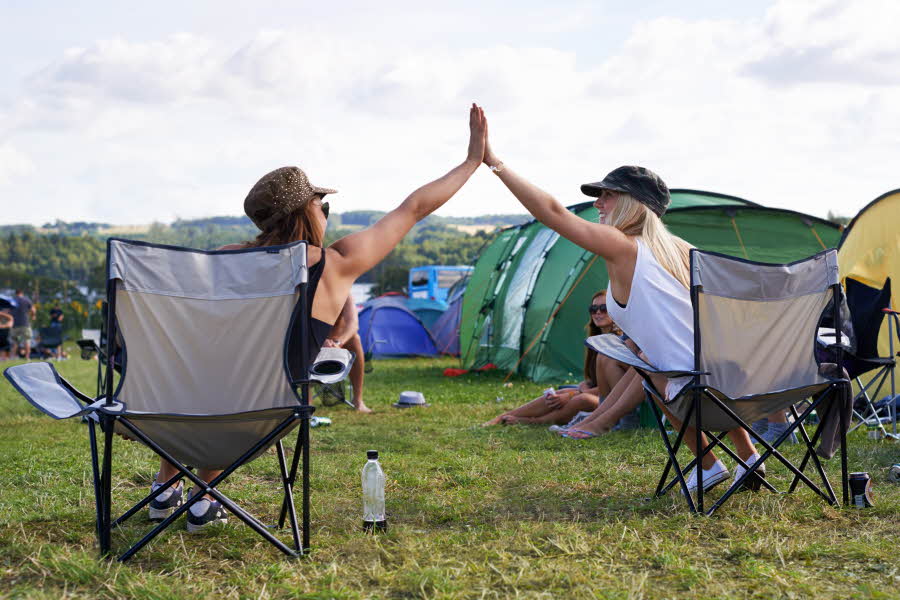 To jenter sitter i hver sin campingstol blant telt og gir hverandre high-five