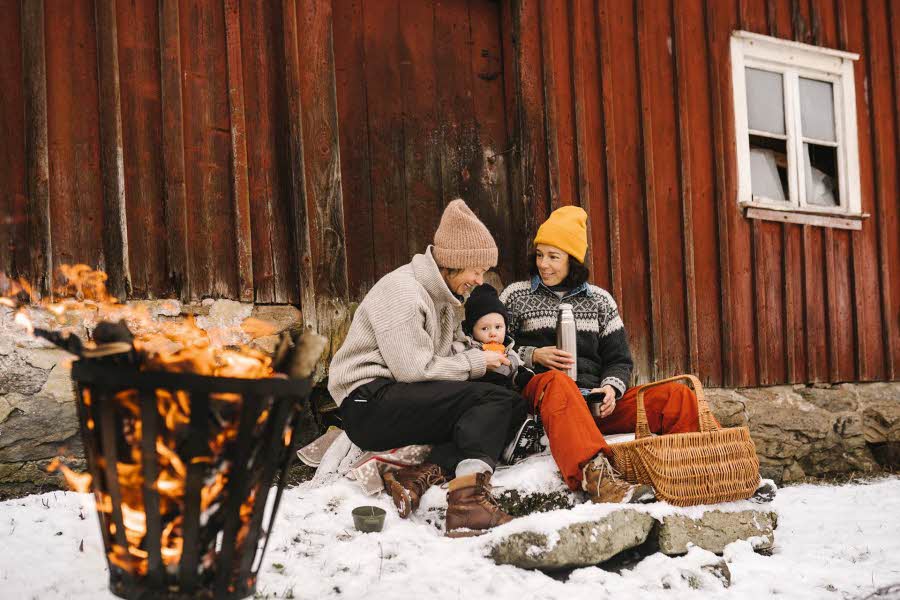 To voksne og et barn sitter ute og smiler inntil en rød vegg i snøen, ved et bål