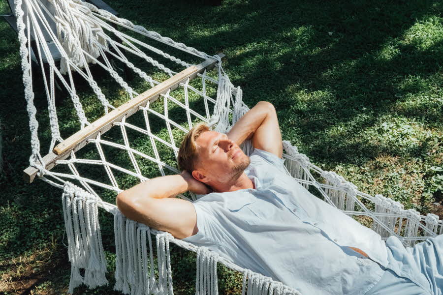 Mann i lyseblå skjorte og shorts slapper av og nyter solen i en hengekøye