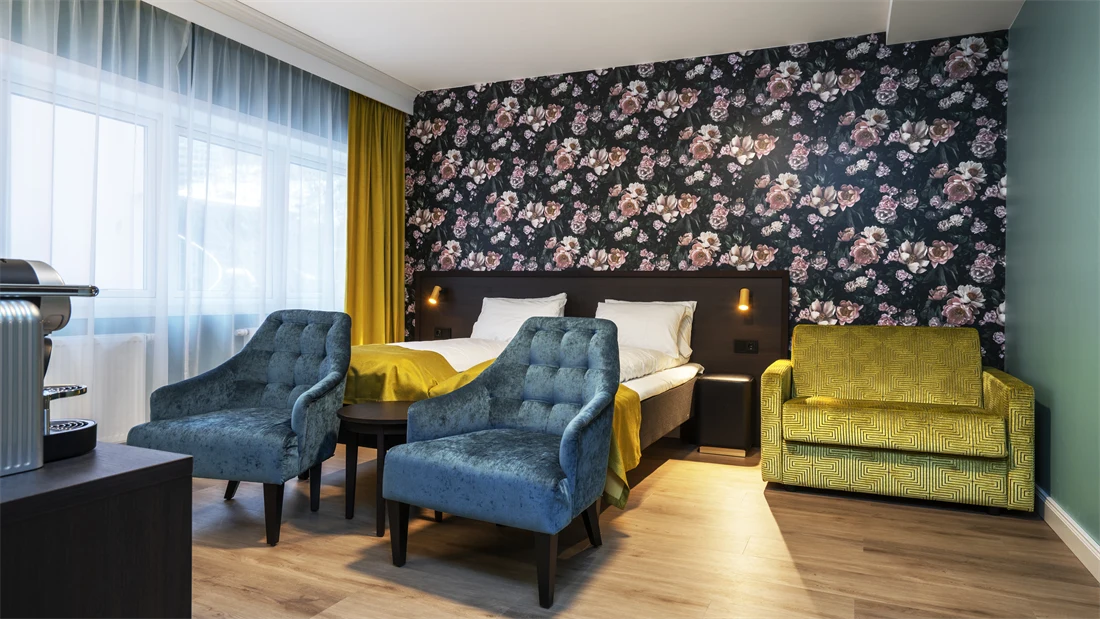  Blomstrete tapet med store lyserosa roser, dobbeltseng, lime grønn sofa, to turkise lenestoler og store vinduer. I superior rom på Thon Hotel Høyers i Skien.