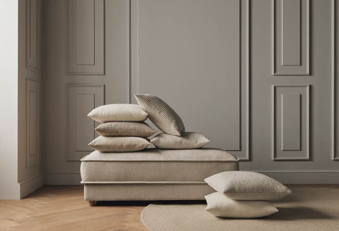 En beige puff med flere puter i nøytrale toner, plassert i et minimalistisk interiør.