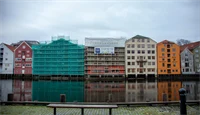 Utsnitt av Kjøpmannsgata som speiler seg i Nidelva, med nr. 15 i midten. I forgrunnen en benk.