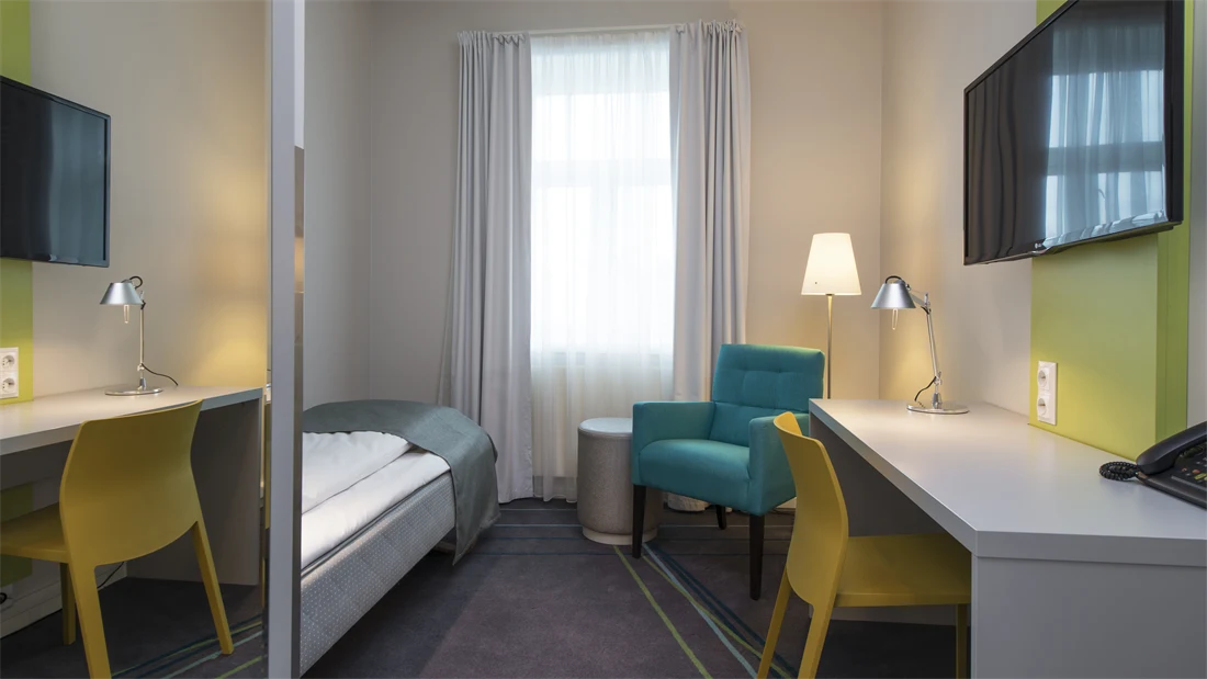 Standard Single Room på Thon Hotel Trondheim. Enkeltseng med hvitt sengetøy og grått sengeteppe. Turkis lenestol, gul kontorstol, hvitt skrivebord, stålampe og vegghengt TV.