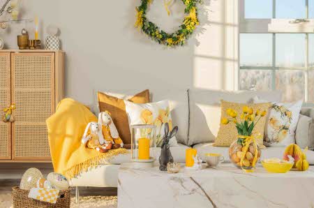En sofa og et stuebord med gule lys, gult pledd, påskeputer, påskeliljer og en kanin på
