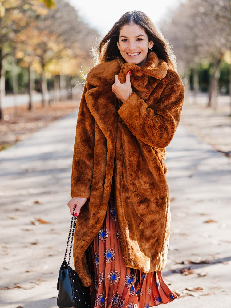 Dame med lang brun fuskepelskåpe smiler til kamera på gaten med høsttrær i bakgrunnen