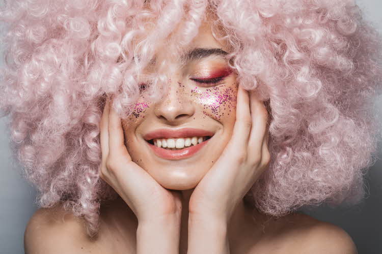 Modell med babyrosa afro og rosa glittersminke holder seg til ansiktet mens hun smiler og lukker øynene