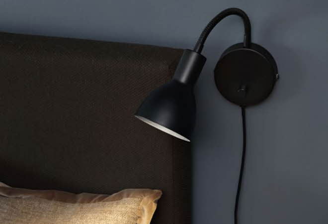 Sort vegglampe med fleksibel arm montert på mørk vegg ved siden av en sengegavl.