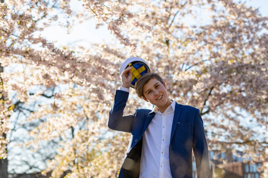En tenåringsgutt i dress og en svensk studentlue. Han står under kirsebærtrær.