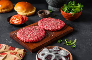 To rå hamburgerpatties på et skjærebrett med ingredienser rundt.