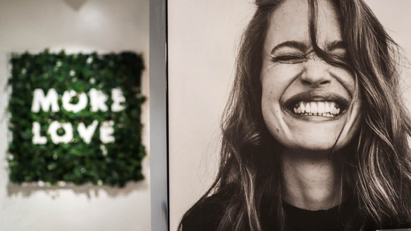 Bilde av dame som smiler, og grønn plantedekor med bokstaver inni "MORE LOVE"