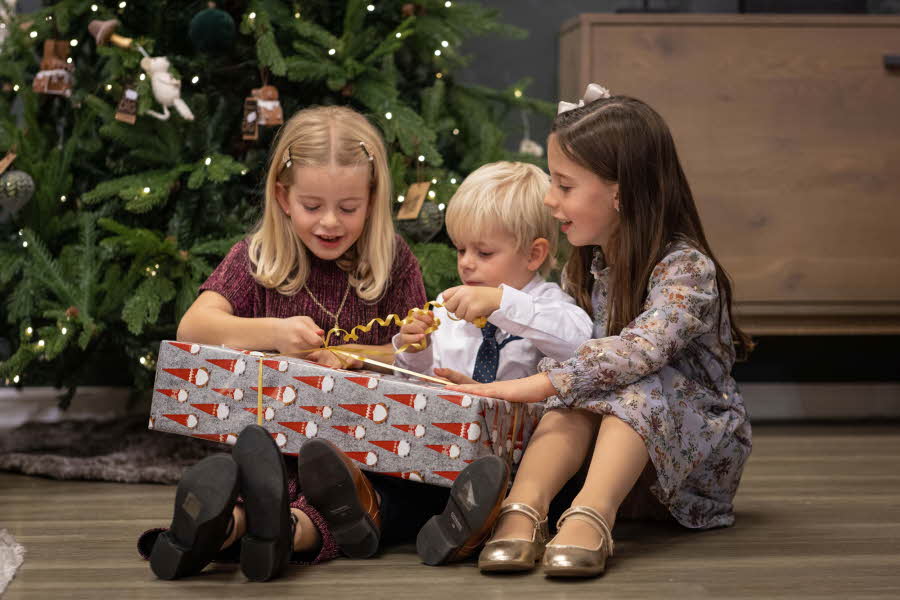 to jenter og en gutt sitter på gulvet foran juletreet og åpner gaver
