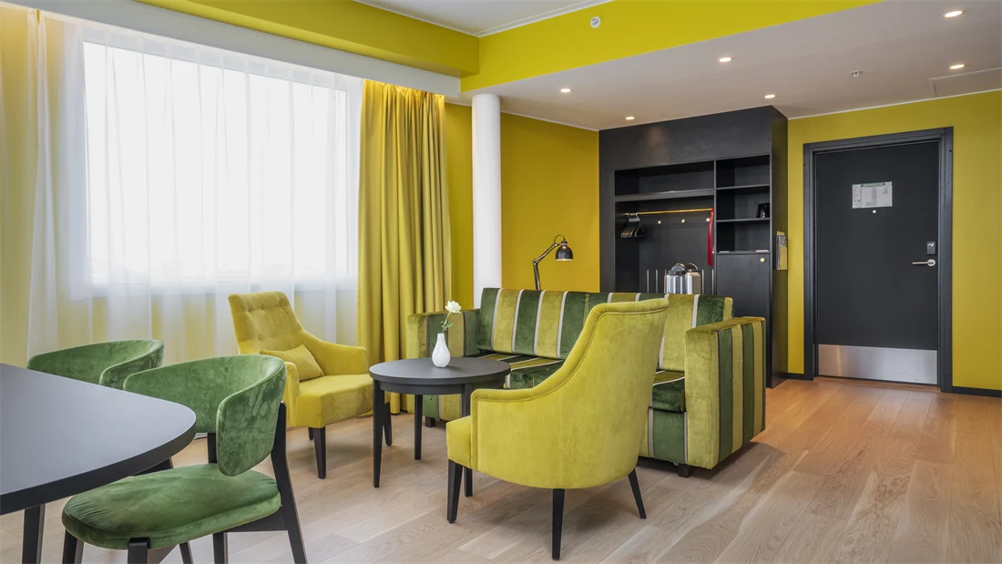 Sittegruppe med gul og grønn stripete sofa, rundt bord, gule stoler, spisegruppe med mørkt rundt bord og grønne stoler, gule vegger og gule gardiner i suite på Thon Hotel Kirkenes