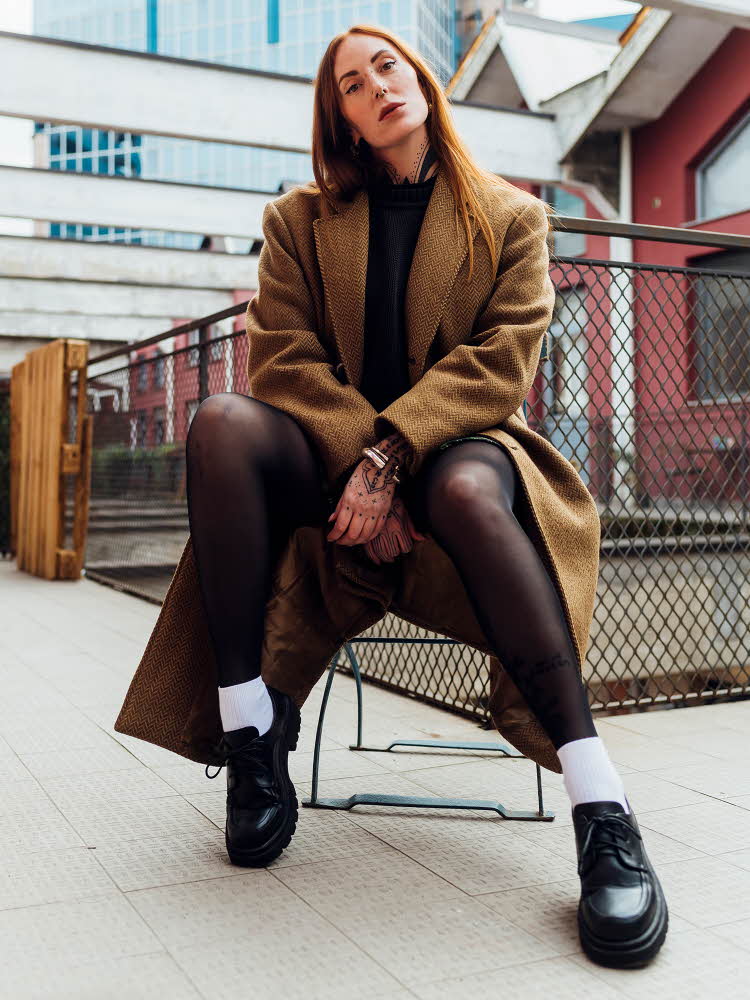 Dame med brun kåpe, sort strømpebukse og sorte snøresko sitter på stol utendørs og poserer for kamera