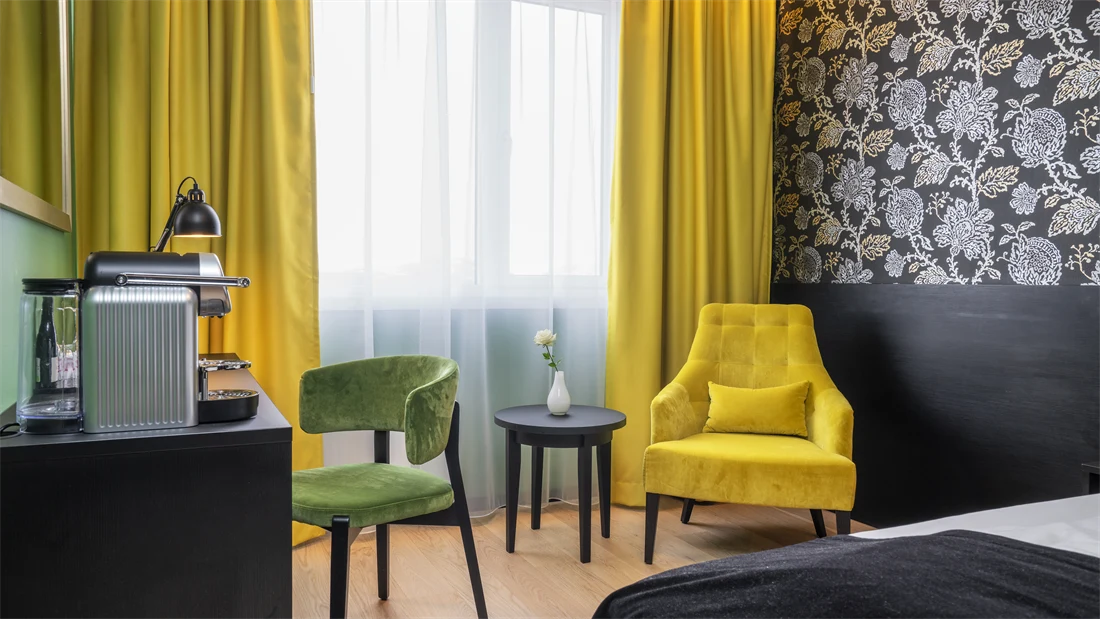 Mørk mønstrete tapet med blomster, gul stol med rundt bord, gule gardiner, skrivebord med kaffemaskin og grønn stol i superior rom på Thon Hotel Kirkenes.