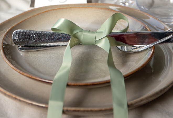 En borddekking med en kniv og gaffel bundet sammen med et lysegrønt bånd på en beige tallerken.