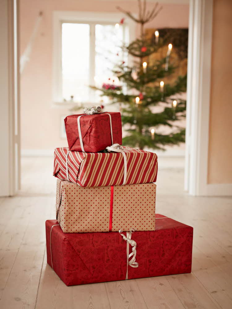 Fire julegaver stablet oppå hverandre i rød innpakning i forskjellige farger, med et pyntet juletre i bakgrunnen