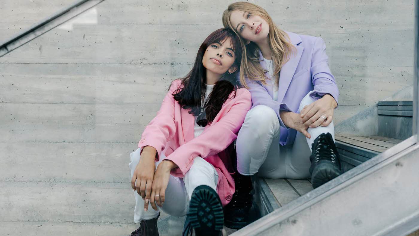to unge damer med pastellfargede, store blazere, sittende i en trapp med hodene inntil hverandre og blikket i kamera