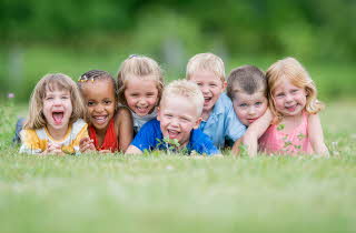 Syv barn som ligger på rekke i gresset, mens de smiler mot kameraet