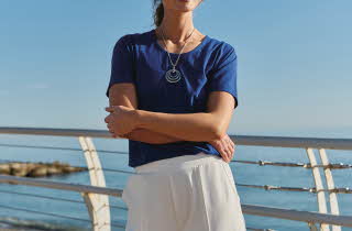 En kvinne står på en bro foran havet, hun har på seg en dyp blå t-skjorte og hvite bukser