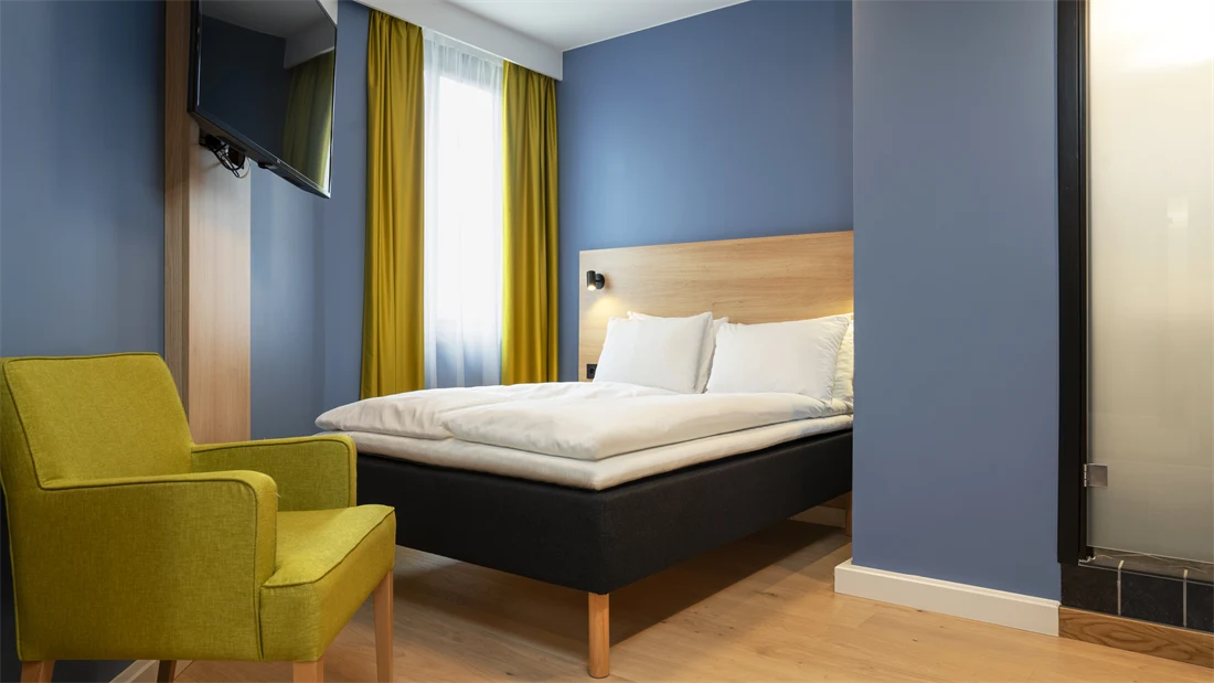 Gul stol, blå vegger, TV og queen seng på standard rom queen på Thon Hotel Astoria.