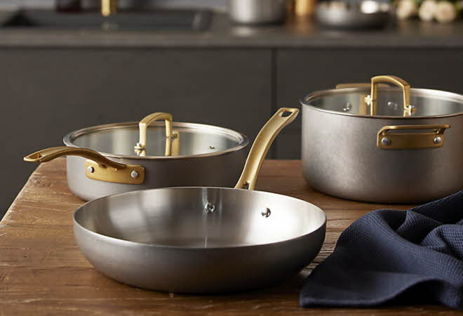 Tre gryter og panner i metall med messinghåndtak på kjøkkenbenk.