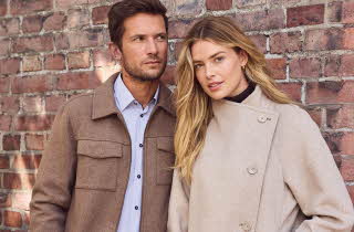 En mann i brun jakke og en kvinne i beige jakke lener seg mot en mursteinsvegg.