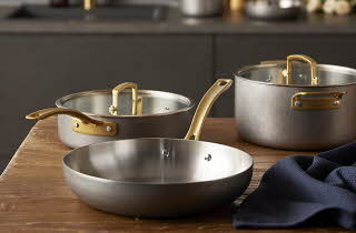 Tre gryter og panner i metall med messinghåndtak på kjøkkenbenk.