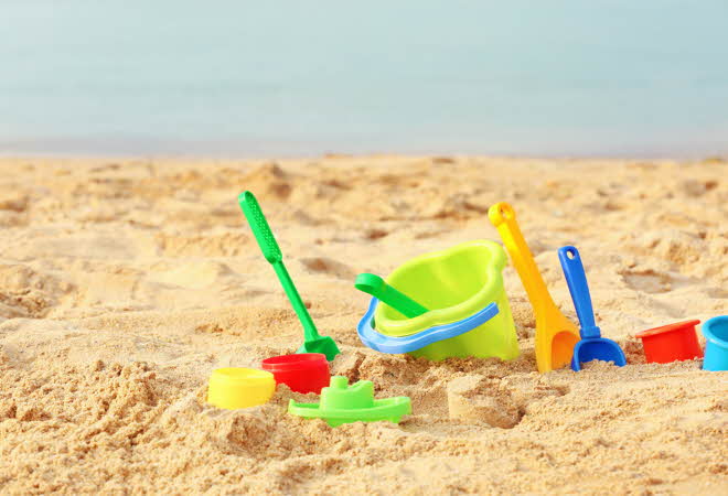 Fargerike plastleker for sand, inkludert bøtter, spader og former, ligger spredt på en strand med gyllen sand og havet i bakgrunnen.