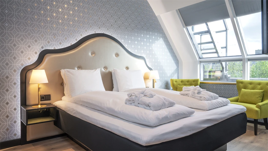 Dobbeltseng med morgenkåper, mønstret sølv tapet, gule stoler og store vinduer i soverom i junior suite på Thon Hotel Cecil