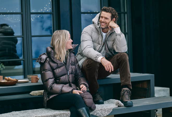 En mann i beige boblejakke og en kvinne i brun boblejakke sitter ute på et bord.