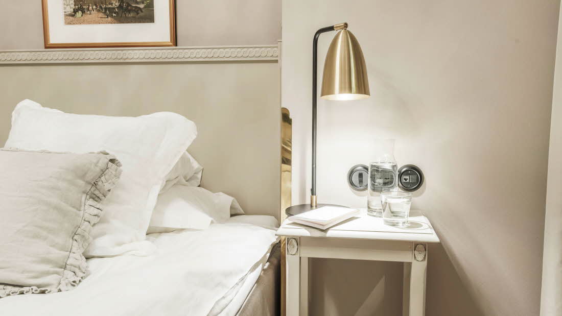 Bild der Ecke eines Betts. Daneben steht ein Nachttisch mit einer Lampe.