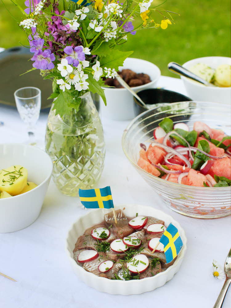 Blond jente smiler til kamera med blomsterkrans på hodet Bord med salat, sild, poteter og fargerik bukett med svenske flagg