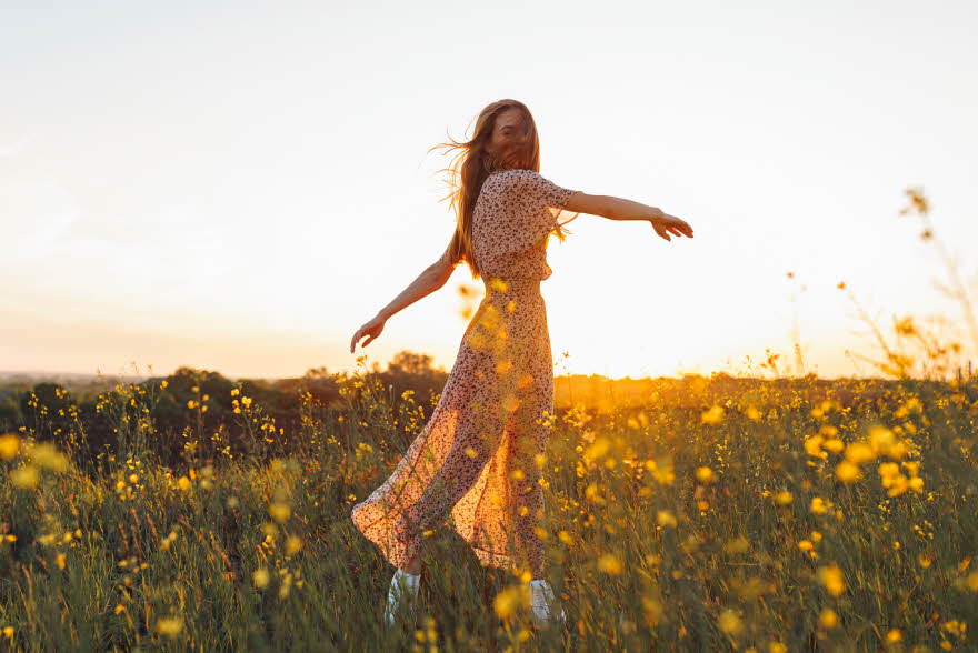 Dame med lang blomstrete kjole danser i gul blomstereng i vårsol