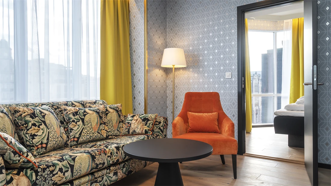 Mønstret fargerik sofa, rundt sort bord, oransje stol, mønstret sølv tapet og gule gardiner i stue på junior suite på Thon Hotel Cecil