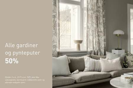 En sofa med pynteputer står foran et vindu med gardiner fra Kid Interiør med teksten "alle gardiner og pynteputer 50%"