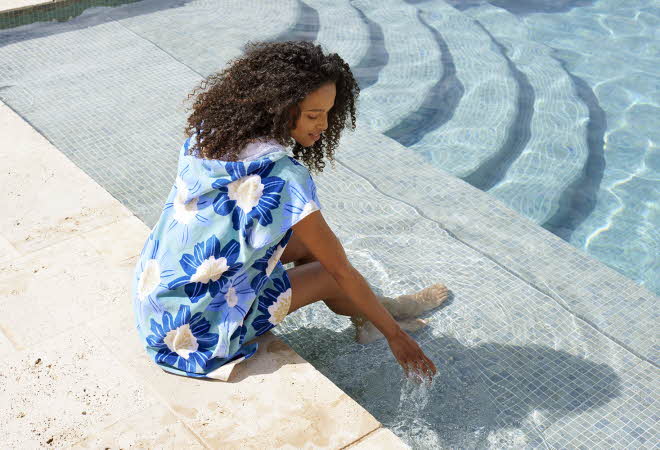 En kvinne som sitter på kanten til et basseng, hun har på seg en blå strandponcho med blå blomster på