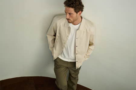 En mann som står i en trapp, og ser ned. Han har på seg en beige jakke med en hvit t-skjorte under, og grønne bukser.