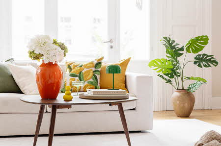 Bilde av en hvit sofa med et stuebord foran. Bordet har en oransj vase med blomster og en liten grønn lampe