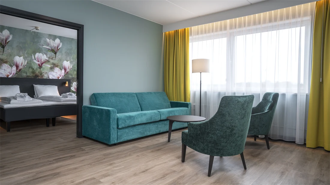 Blomstertapet, dobbeltseng, grønn sofa, stue, salongbord, to grønne lenestoler, vindu, gule gardiner, turkis/grønn vegg
