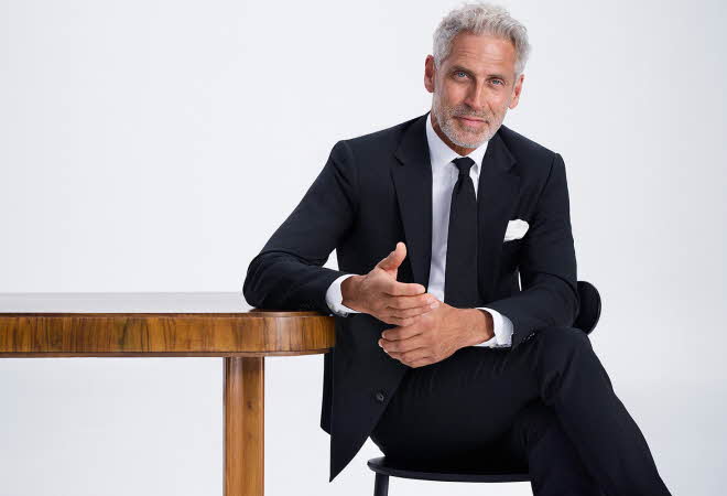 Mann i svart dress og hvit skjorte sittende ved et bord.