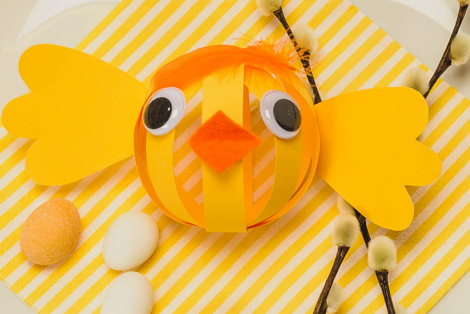 Papir-påskekylling laget av gult papir med oransje nebb og fjær til hår på en tallerken