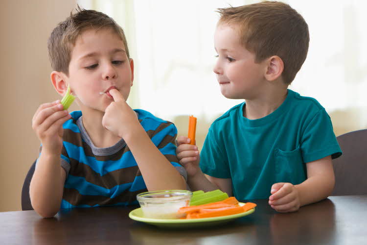 To gutter spiser grønnsaker og dipp ved et bord.